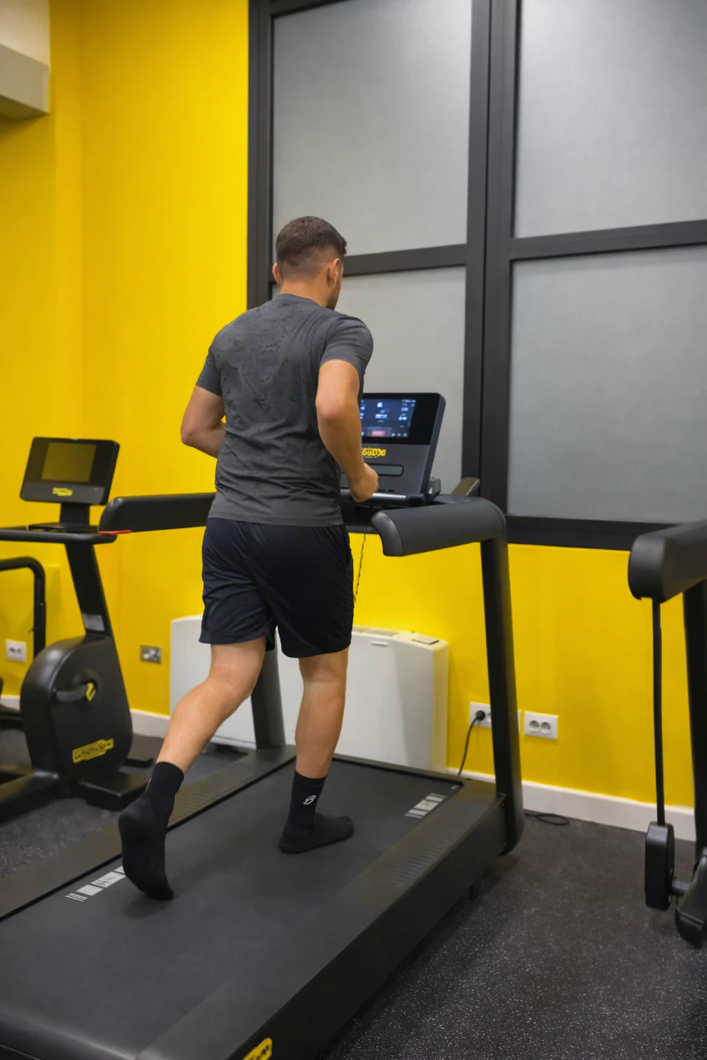 Persona ripresa di spalle che cammina su un tapis roulant in una palestra dalle pareti gialle